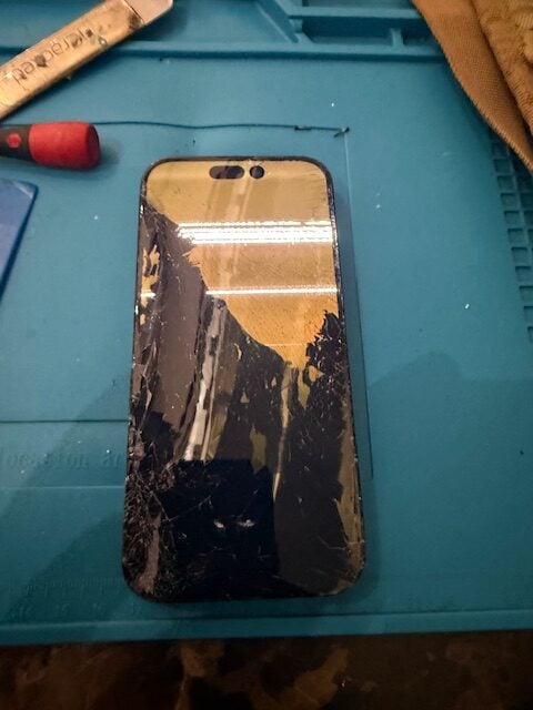 Severely cracked iPhone 15 Pro screen on a blue repair mat before replacement at Heathrow