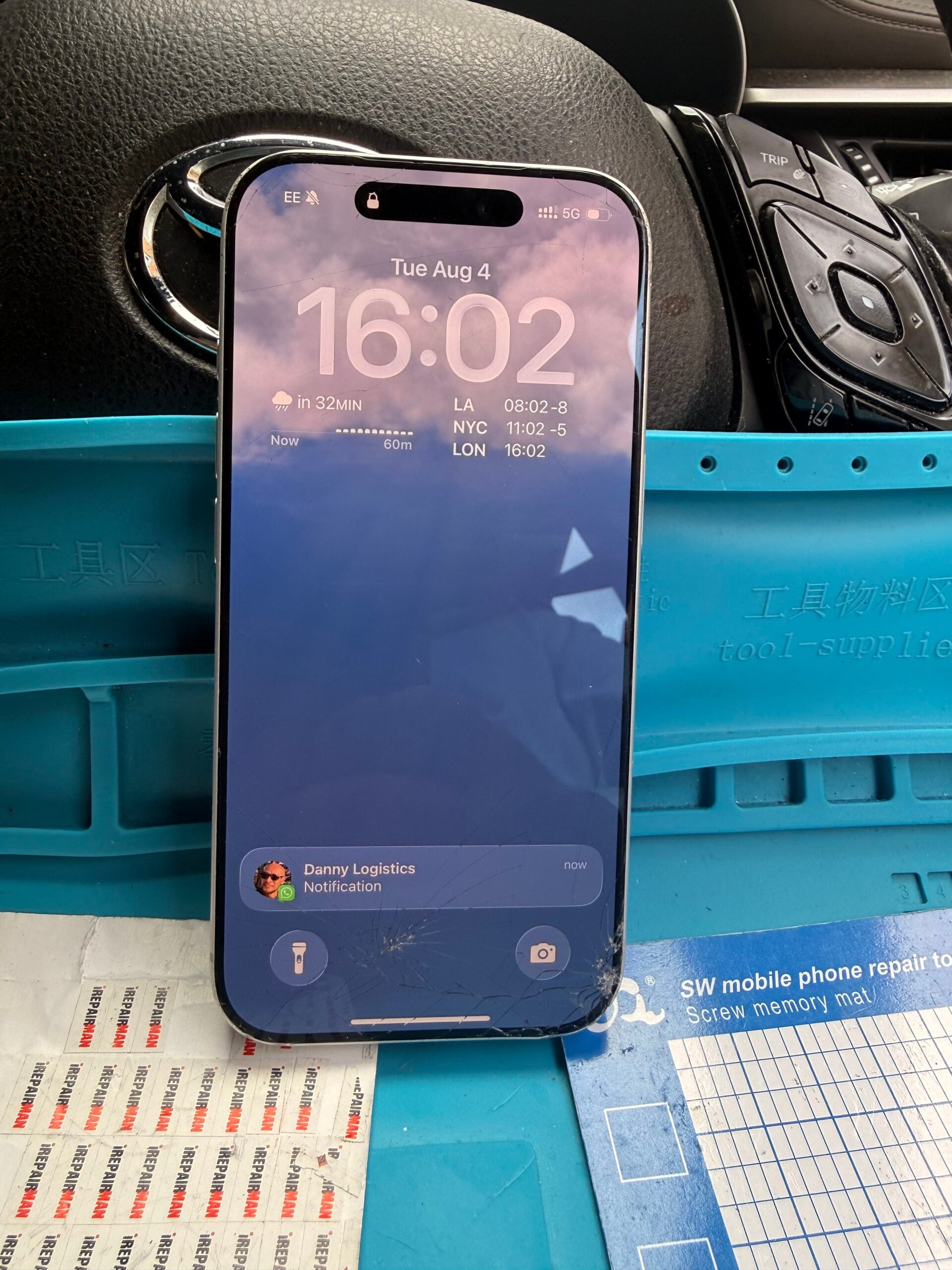 iPhone 17 Pro showing its lock screen with cracked glass across the lower edge on a turquoise repair mat.