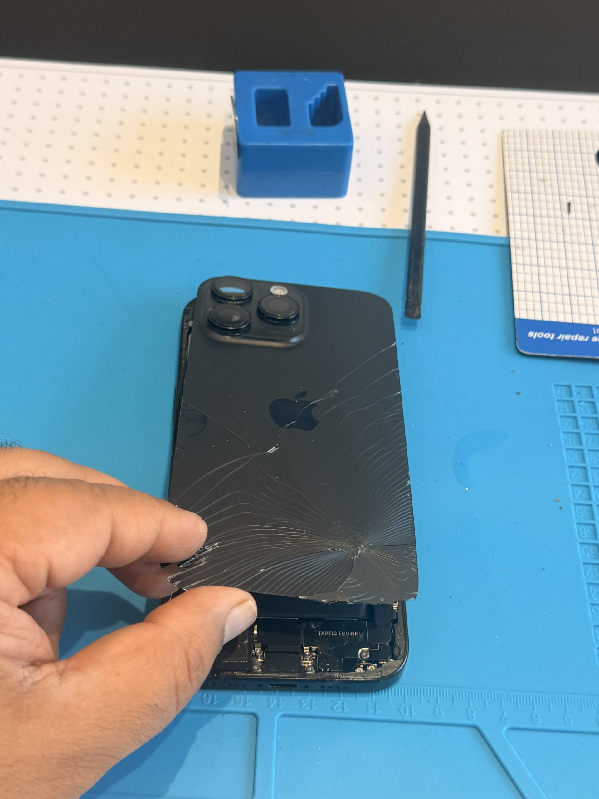A technician lifts the shattered iPhone 16 Pro Max rear panel, exposing the lower internal components on a blue repair mat.
