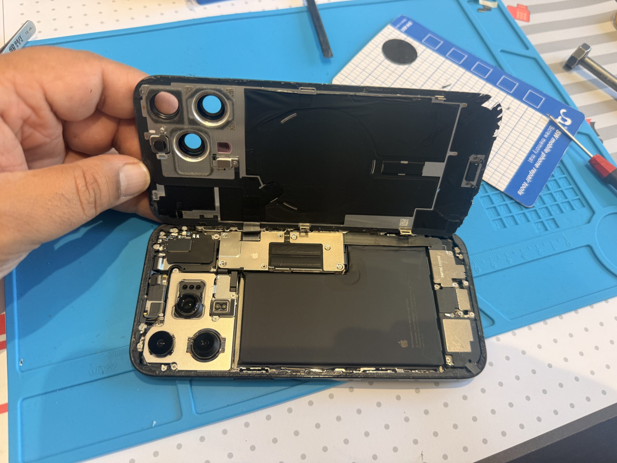 An opened iPhone 16 Pro Max with its rear panel raised above the housing, revealing the battery, cameras and internal components.