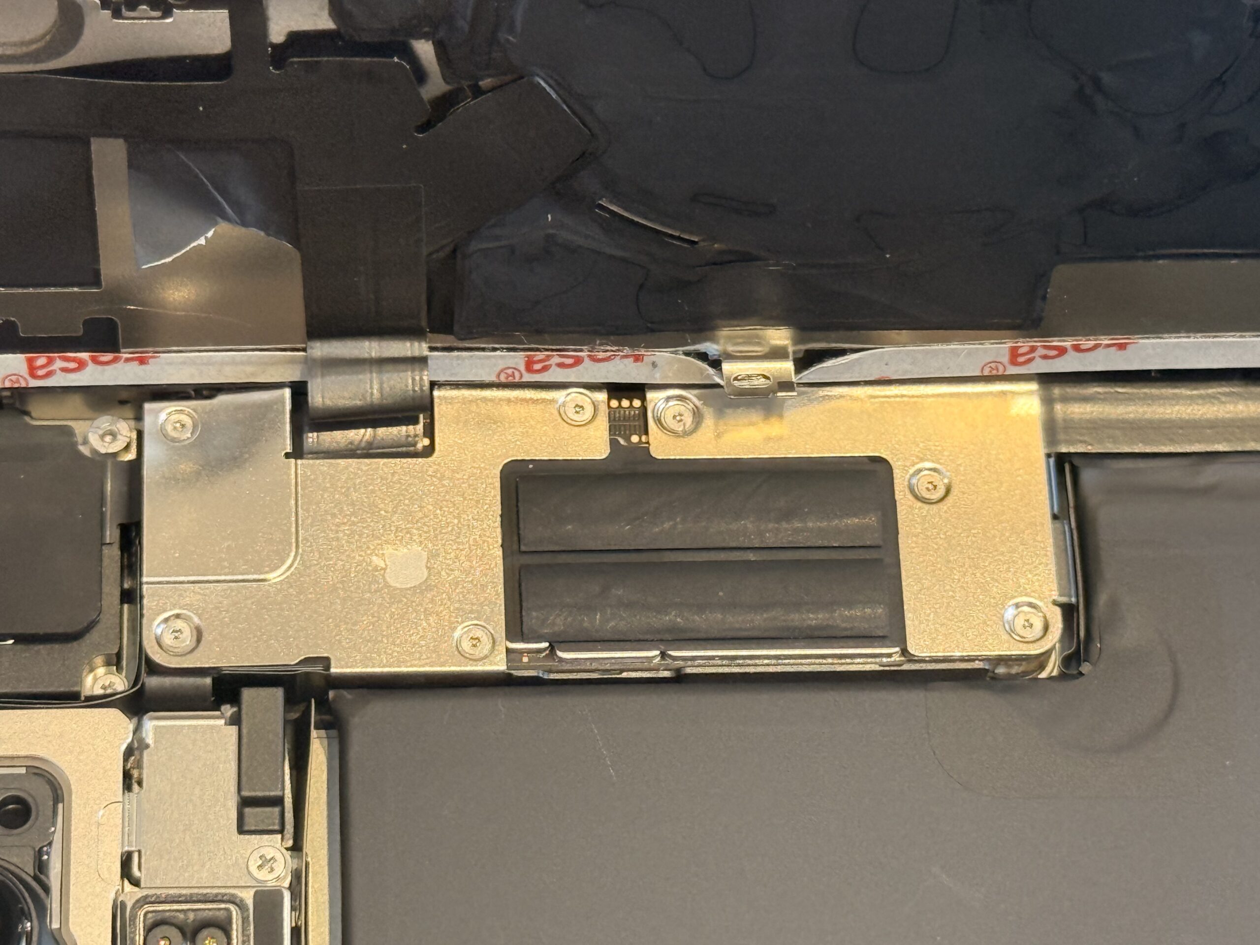 Close-up of the opened iPhone 16 Pro Max showing a rear-panel ribbon cable, metal brackets and screws above the battery.