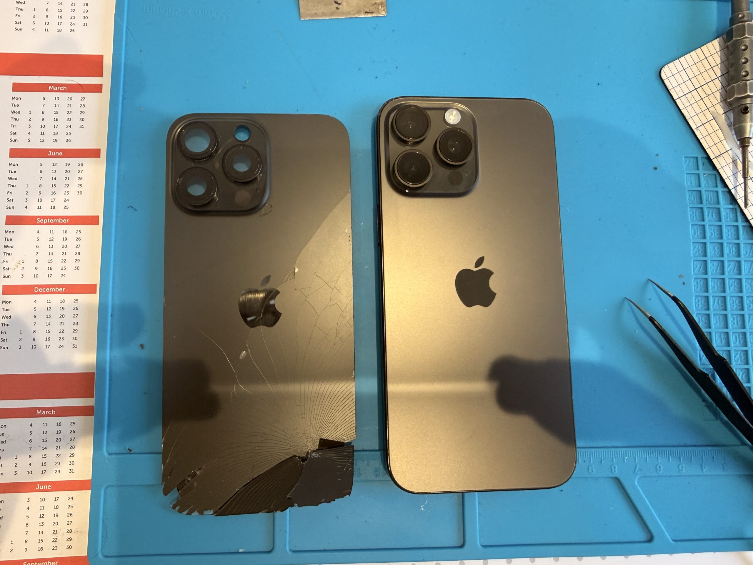 The removed cracked iPhone 16 Pro Max back panel lies beside the repaired phone with intact rear glass on a blue repair mat.